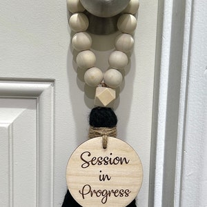 May include: A wooden door hanger with the text "Session in Progress". The door hanger has a wooden bead garland, a black tassel, and a wooden disc with the text in a script font. The door is white.