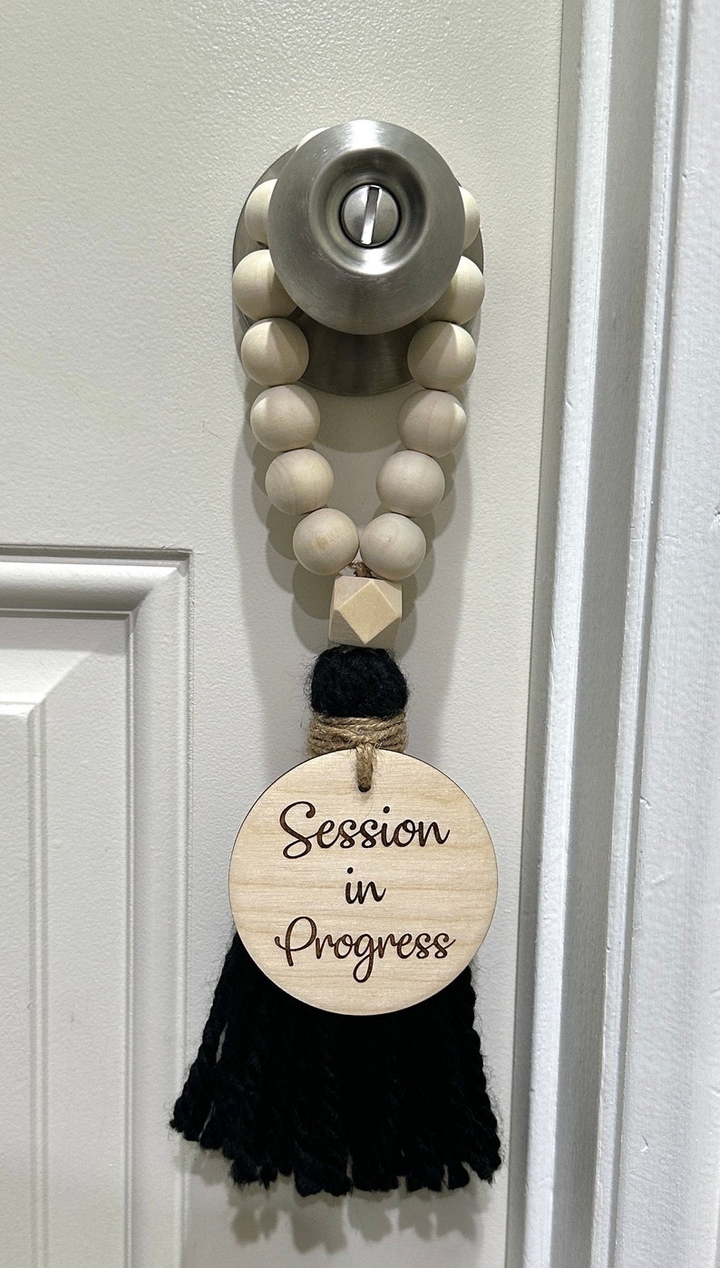 May include: A wooden door hanger with the text "Session in Progress". The door hanger has a wooden bead garland, a black tassel, and a wooden disc with the text in a script font. The door is white.
