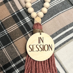 May include: A wooden bead garland with a geometric wooden accent and a tassel. The tassel is made of a dusty rose yarn and is attached to a wooden disc that reads "IN SESSION".