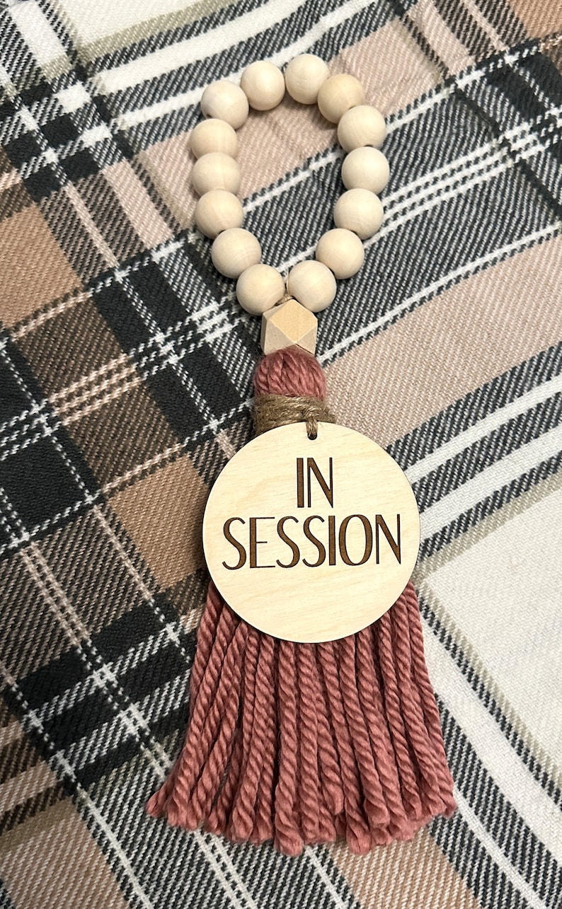 May include: A wooden bead garland with a geometric wooden accent and a tassel. The tassel is made of a dusty rose yarn and is attached to a wooden disc that reads "IN SESSION".