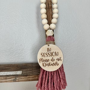 May include: A wooden sign with the text "IN SESSION Please do not Disturb" hangs from a string of wooden beads. The sign is attached to a pink tassel. The beads are light-colored and round.