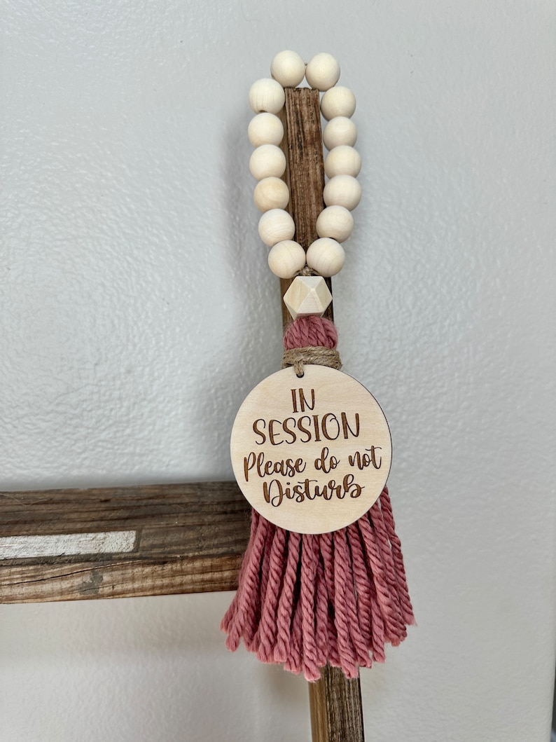 May include: A wooden sign with the text "IN SESSION Please do not Disturb" hangs from a string of wooden beads. The sign is attached to a pink tassel. The beads are light-colored and round.