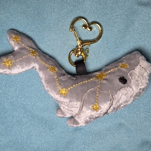 May include: A plush gray whale keychain with gold embroidered constellation details. The whale has a black eye and a gold heart-shaped clasp. The keychain is on a blue background.
