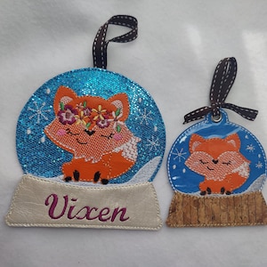 May include: Two blue and white snow globe ornaments with a fox inside. The larger ornament has the name "Vixen" on the base.