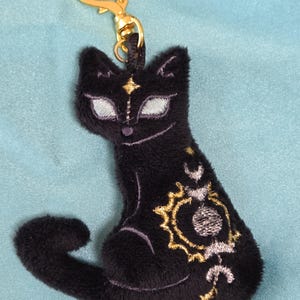 May include: Black plush cat keychain with gold-colored hardware. The cat has embroidered details, including a star on its forehead and celestial designs on its side. The keychain has a heart-shaped clasp.