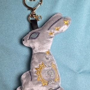 May include: A gray plush rabbit keychain with gold-colored accents. The rabbit has embroidered details, including stars, a crescent moon, and a sun design. The keychain has a gold-colored heart-shaped clasp and a black strap.