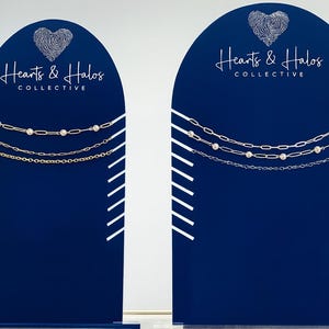 May include: Two navy blue display stands with white diagonal stripes and the text "Hearts & Halos COLLECTIVE". Each stand features three silver necklaces with various chain styles and embellishments, including pearls and round gems.
