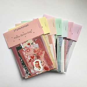 May include: A collection of assorted paper scraps and stickers in a clear plastic sleeve, topped with a pink paper header labeled paper & poem mini grab bag. The contents feature various floral, heart, and bird patterns in pink, red, and green.