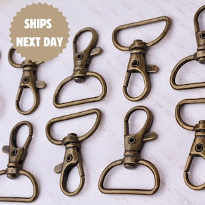 Puede incluir: Primer plano de varios cierres giratorios de color bronce antiguo con anillas en D. Los cierres están dispuestos sobre una superficie blanca. El texto "SHIPS NEXT DAY" está en forma de estrella marrón en la esquina superior izquierda. Ideal para manualidades.