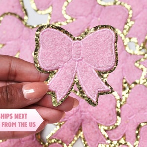 Puede incluir: Un parche en forma de lazo rosa claro con un borde de purpurina dorada. El parche tiene un aspecto texturizado y está rodeado de otros parches similares. El texto "SHIPS NEXT DAY FROM THE US" es visible.