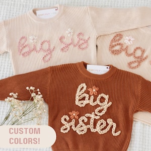 May include: Two knitted sweaters with the words "Big Sis" and "Big Sister" embroidered on them. The sweaters are in different colors, one is cream and the other is brown. The sweaters are laid out on a white surface.