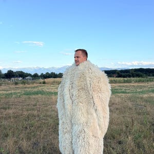 Könnte beinhalten: Ein langer, cremefarbener Mantel aus dicker, zotteliger Schaffellwolle. Das Kleidungsstück verfügt über einen hohen, voluminösen Kragen und einen dichten, strukturierten Flor, der die gesamte Außenseite bedeckt. Der Mantel reicht bis zur Wadenmitte.