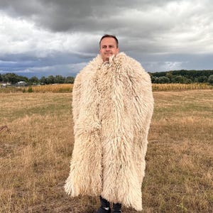Op de afbeelding: Een lange, zware jas gemaakt van dikke, crèmekleurige schapenvacht met een ruige, getextureerde wollen buitenkant. Het kledingstuk heeft een hoge kraag en een losse, oversized pasvorm die tot op de knieën valt en het bovenlichaam volledig bedekt.