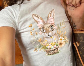 May include: White t-shirt with a graphic of a bunny wearing glasses and a pink bow, holding a basket of Easter eggs.