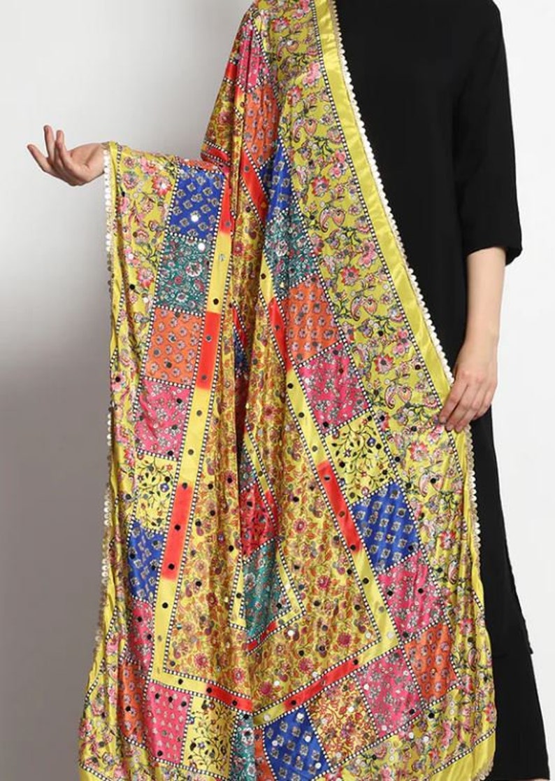 May include: A yellow silk dupatta with a patchwork design featuring colorful squares and floral patterns. The dupatta has a white border with small pearl-like embellishments and is adorned with small round mirrors.