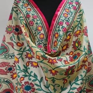 K&ouml;nnte beinhalten: Cremefarbener bestickter Schal mit einem lebendigen Blumenmuster. Das Design zeigt rosa, gelbe und t&uuml;rkisfarbene Blumen mit gr&uuml;nen Bl&auml;ttern, die mit einer kontrastierenden Naht umrandet sind. Ein leuchtend pinkfarbener Rand rahmt die Kanten ein und erzeugt einen auff&auml;lligen visuellen Kontrast.