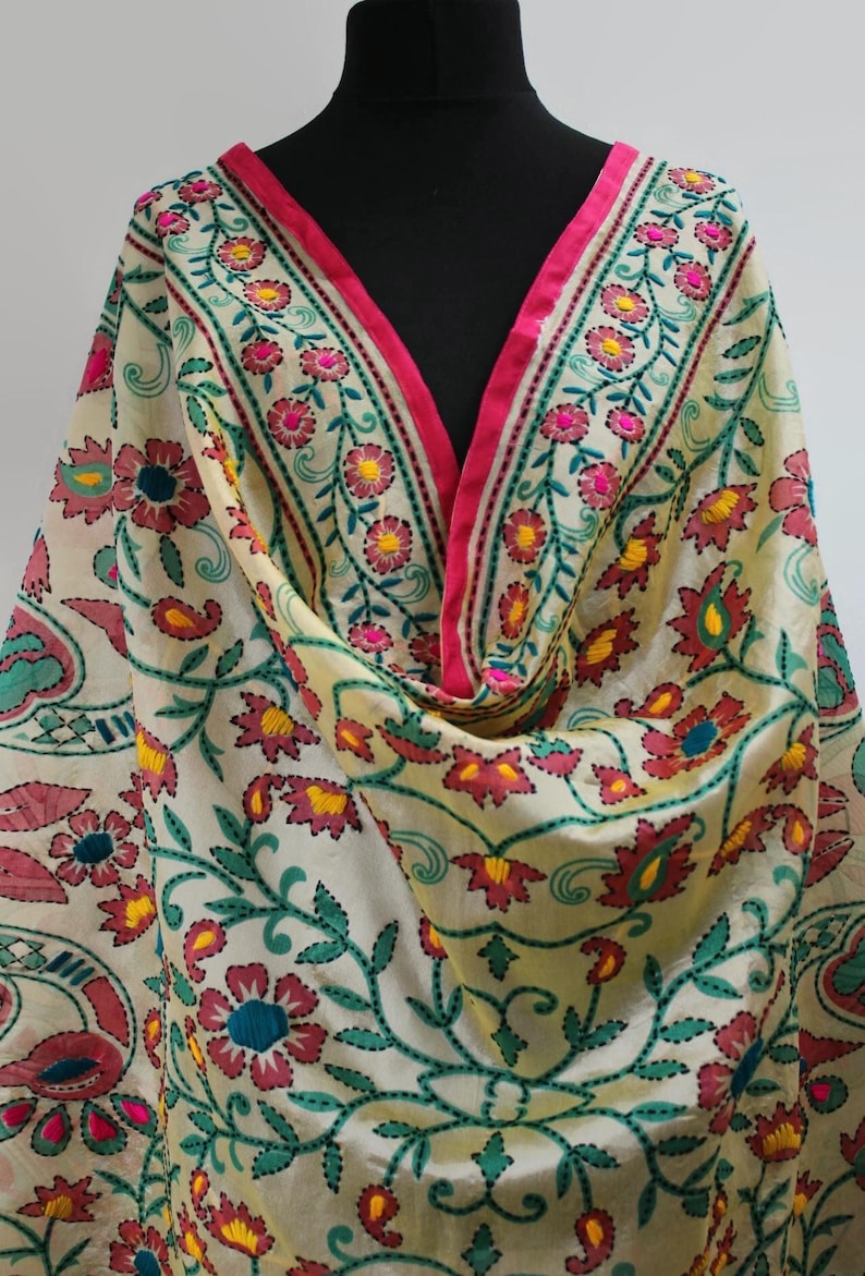 K&ouml;nnte beinhalten: Cremefarbener bestickter Schal mit einem lebendigen Blumenmuster. Das Design zeigt rosa, gelbe und t&uuml;rkisfarbene Blumen mit gr&uuml;nen Bl&auml;ttern, die mit einer kontrastierenden Naht umrandet sind. Ein leuchtend pinkfarbener Rand rahmt die Kanten ein und erzeugt einen auff&auml;lligen visuellen Kontrast.