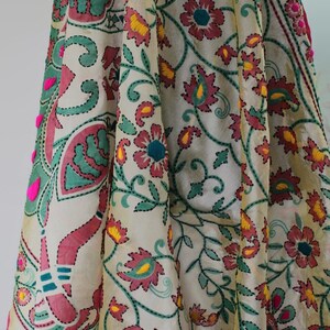 K&ouml;nnte beinhalten: Cremefarbener Stoff mit aufwendiger Blumenstickerei in Rosa-, T&uuml;rkis- und Gelbt&ouml;nen. Das Design weist detaillierte N&auml;hte und dekorative Elemente auf, darunter kleine rosa Pompons am Rand. Der Stoff ist drapiert und zeigt das Muster.