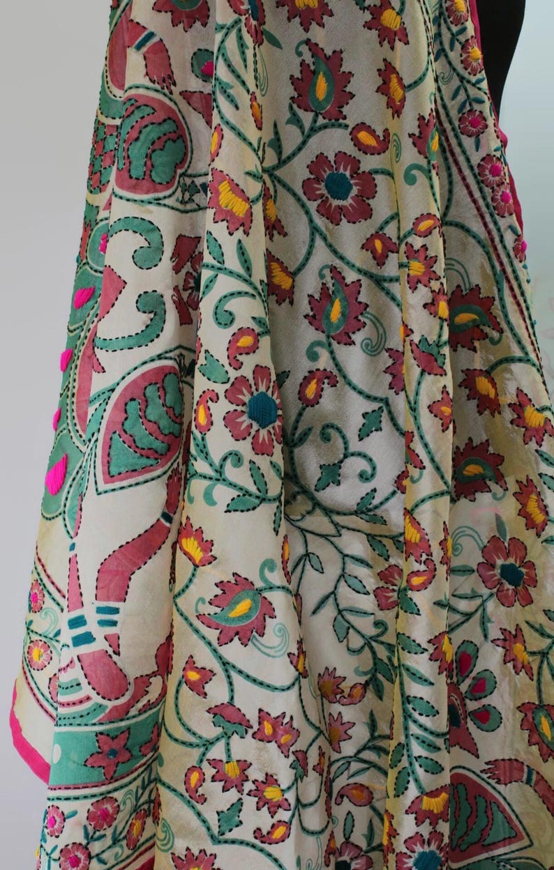 K&ouml;nnte beinhalten: Cremefarbener Stoff mit aufwendiger Blumenstickerei in Rosa-, T&uuml;rkis- und Gelbt&ouml;nen. Das Design weist detaillierte N&auml;hte und dekorative Elemente auf, darunter kleine rosa Pompons am Rand. Der Stoff ist drapiert und zeigt das Muster.