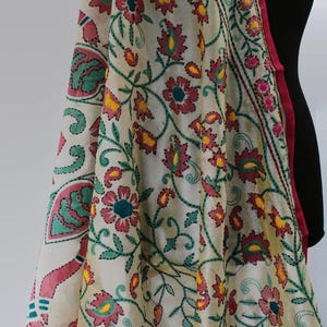 K&ouml;nnte beinhalten: Cremefarbener bestickter Schal mit einem Blumenmuster in Rosa-, Gelb- und Gr&uuml;nt&ouml;nen. Der Schal hat einen rosa Rand und ist &uuml;ber einer schwarzen Schaufensterpuppe drapiert. Die Stickerei ist detailliert und aufwendig.