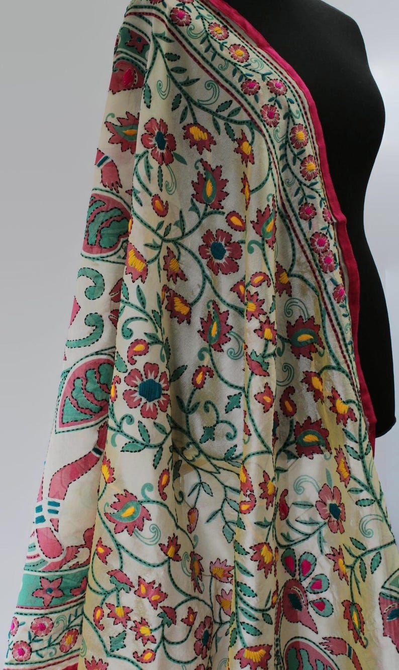 K&ouml;nnte beinhalten: Cremefarbener bestickter Schal mit einem Blumenmuster in Rosa-, Gelb- und Gr&uuml;nt&ouml;nen. Der Schal hat einen rosa Rand und ist &uuml;ber einer schwarzen Schaufensterpuppe drapiert. Die Stickerei ist detailliert und aufwendig.