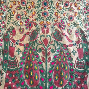 K&ouml;nnte beinhalten: Dekoratives Textil mit detailliertem Stickdesign. Das Kunstwerk zeigt zwei Figuren, die ein zentrales Pfauenmotiv flankieren, mit floralen und Blattmustern in Rosa, Gr&uuml;n, Gelb und Blau. Der Rand ist leuchtend rosa.