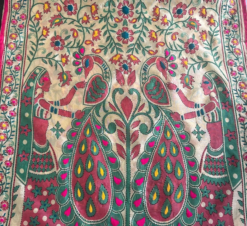 K&ouml;nnte beinhalten: Dekoratives Textil mit detailliertem Stickdesign. Das Kunstwerk zeigt zwei Figuren, die ein zentrales Pfauenmotiv flankieren, mit floralen und Blattmustern in Rosa, Gr&uuml;n, Gelb und Blau. Der Rand ist leuchtend rosa.