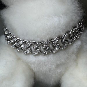 Puede incluir: Un collar de perro de cadena plateada con diamantes de imitación transparentes. El collar es grueso y tiene un diseño de eslabones gruesos.