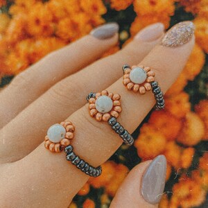 May include: Three beaded rings with a white stone center and copper colored beads. The rings are on a hand with a light orange background.