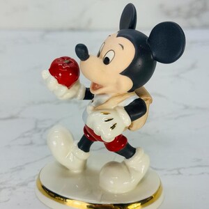 May include: A porcelain figurine of Mickey Mouse holding a red apple. He is wearing a red and white shirt, red shorts, and a brown backpack. The figurine is standing on a white base with a gold trim.