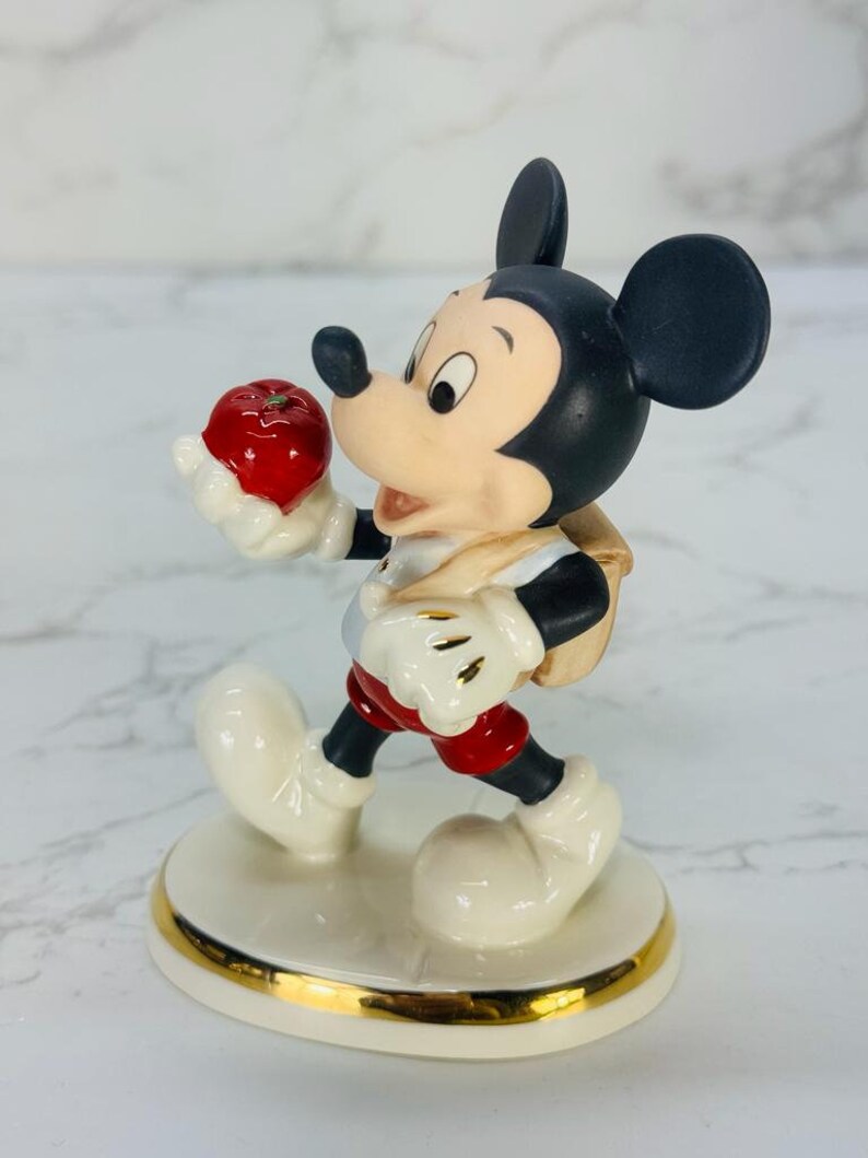 May include: A porcelain figurine of Mickey Mouse holding a red apple. He is wearing a red and white shirt, red shorts, and a brown backpack. The figurine is standing on a white base with a gold trim.
