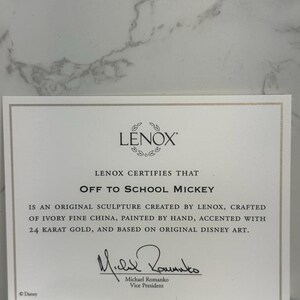 May include: A white certificate with gold trim, certifying that the Off to School Mickey sculpture is an original creation by Lenox. It is crafted from ivory fine china, hand-painted, accented with 24 karat gold, and based on original Disney art. The certificate is signed by Michael Romanko, Vice President.
