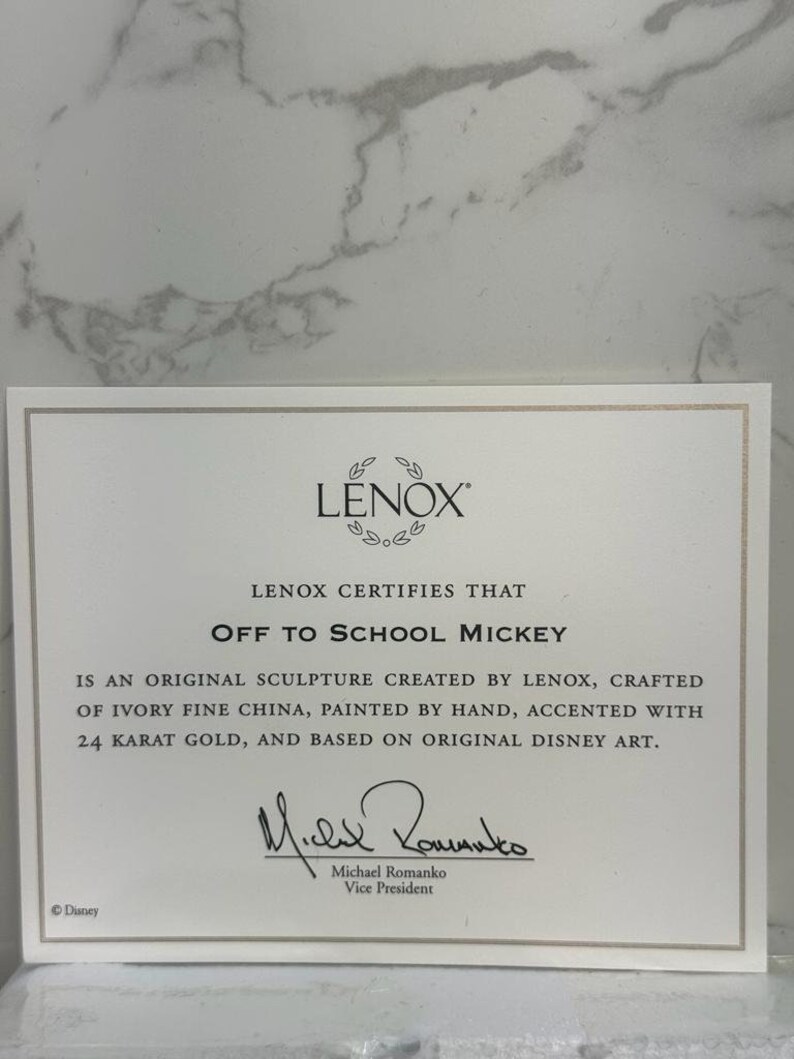 May include: A white certificate with gold trim, certifying that the Off to School Mickey sculpture is an original creation by Lenox. It is crafted from ivory fine china, hand-painted, accented with 24 karat gold, and based on original Disney art. The certificate is signed by Michael Romanko, Vice President.