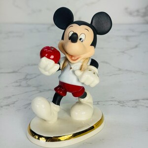 May include: A porcelain figurine of Mickey Mouse holding a red apple. He is wearing a red and white shirt, red shorts, and white shoes. The figurine is standing on a white base with a gold trim.