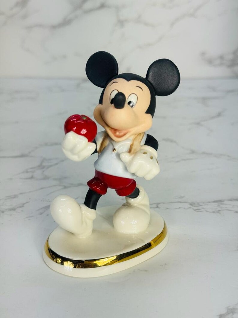 May include: A porcelain figurine of Mickey Mouse holding a red apple. He is wearing a red and white shirt, red shorts, and white shoes. The figurine is standing on a white base with a gold trim.