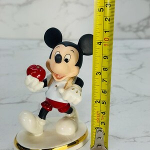 May include: A porcelain figurine of Mickey Mouse holding a red apple. He is wearing a white shirt with a red collar, red shorts, and black shoes. The figurine is standing on a white base with a gold trim. It is approximately 4 inches tall.