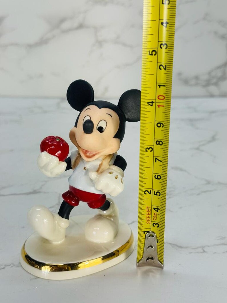 May include: A porcelain figurine of Mickey Mouse holding a red apple. He is wearing a white shirt with a red collar, red shorts, and black shoes. The figurine is standing on a white base with a gold trim. It is approximately 4 inches tall.