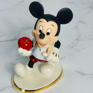 May include: A porcelain figurine of Mickey Mouse holding a red apple. He is wearing a white shirt with a red collar, red shorts, and black shoes. The figurine is standing on a white oval base with a gold trim.