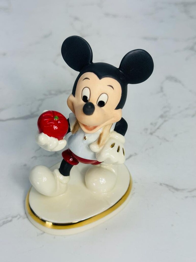 May include: A porcelain figurine of Mickey Mouse holding a red apple. He is wearing a white shirt with a red collar, red shorts, and black shoes. The figurine is standing on a white oval base with a gold trim.