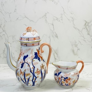 May include: A white porcelain teapot and creamer set with a floral pattern in shades of blue, orange, and gold. The teapot has a pink handle and a floral lid with a small pink frog finial. The creamer has a pink handle.
