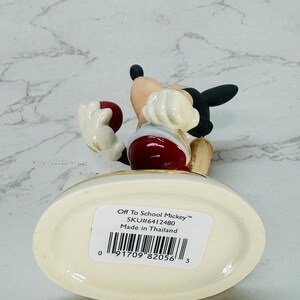 May include: A ceramic figurine of Mickey Mouse dressed in red and white, holding a red ball. The figurine is standing on a white base with a gold trim. The base has a label that reads "Off To School Mickey&trade; SKU#6412480 Made in Thailand" and a barcode.