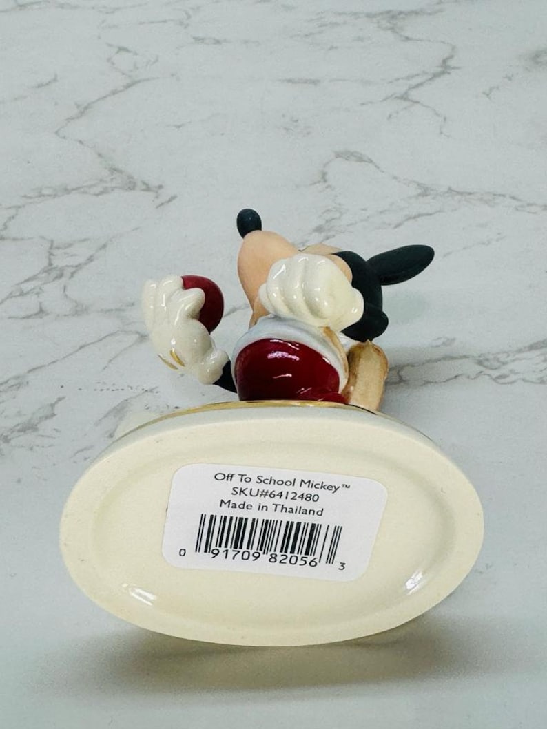 May include: A ceramic figurine of Mickey Mouse dressed in red and white, holding a red ball. The figurine is standing on a white base with a gold trim. The base has a label that reads "Off To School Mickey&trade; SKU#6412480 Made in Thailand" and a barcode.
