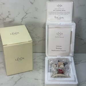 May include: A white Lenox figurine of Mickey Mouse wearing a red and white baseball uniform, holding a baseball bat and a baseball. The figurine is in a white box with a certificate of authenticity that reads "Lenox certifies that Off to School Mickey is an original sculpture crafted by Lenox, crafted of ivory fine china, painted by hand, accented with 14 karat gold, and based on original Disney art."