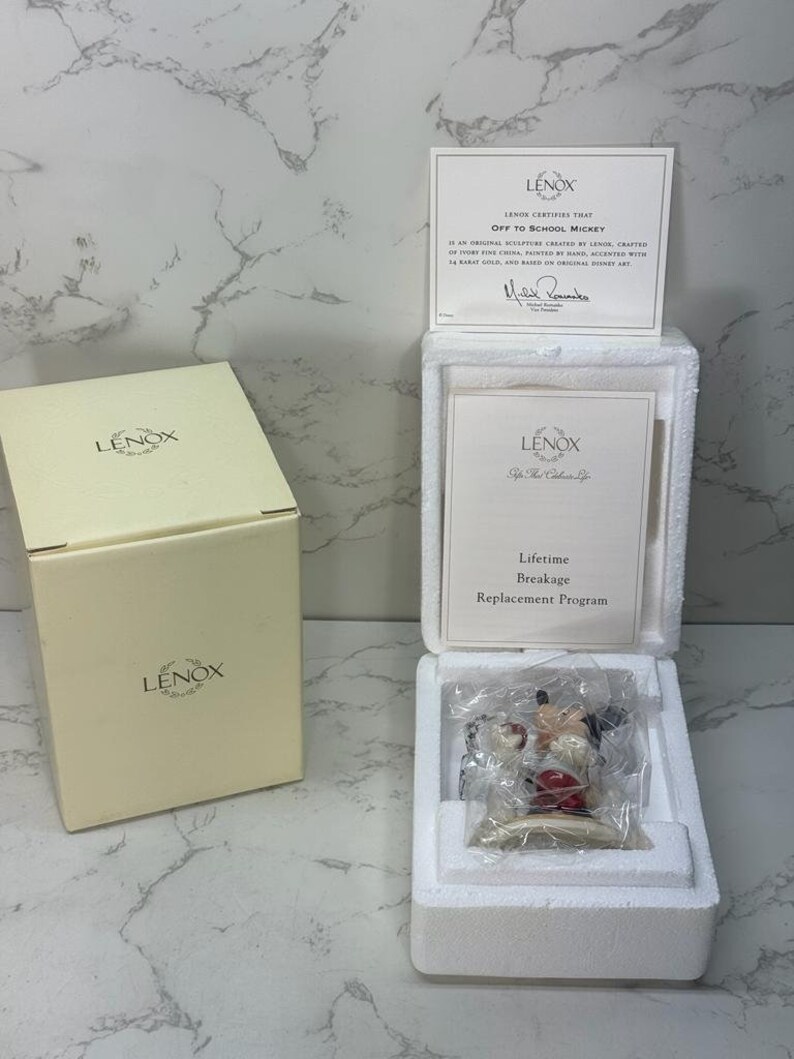 May include: A white Lenox figurine of Mickey Mouse wearing a red and white baseball uniform, holding a baseball bat and a baseball. The figurine is in a white box with a certificate of authenticity that reads "Lenox certifies that Off to School Mickey is an original sculpture crafted by Lenox, crafted of ivory fine china, painted by hand, accented with 14 karat gold, and based on original Disney art."