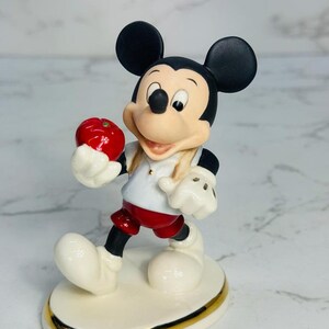 May include: A porcelain figurine of Mickey Mouse holding a red apple. He is wearing a white shirt with a red collar, red shorts, and white gloves. The figurine is standing on a white base with a gold trim.