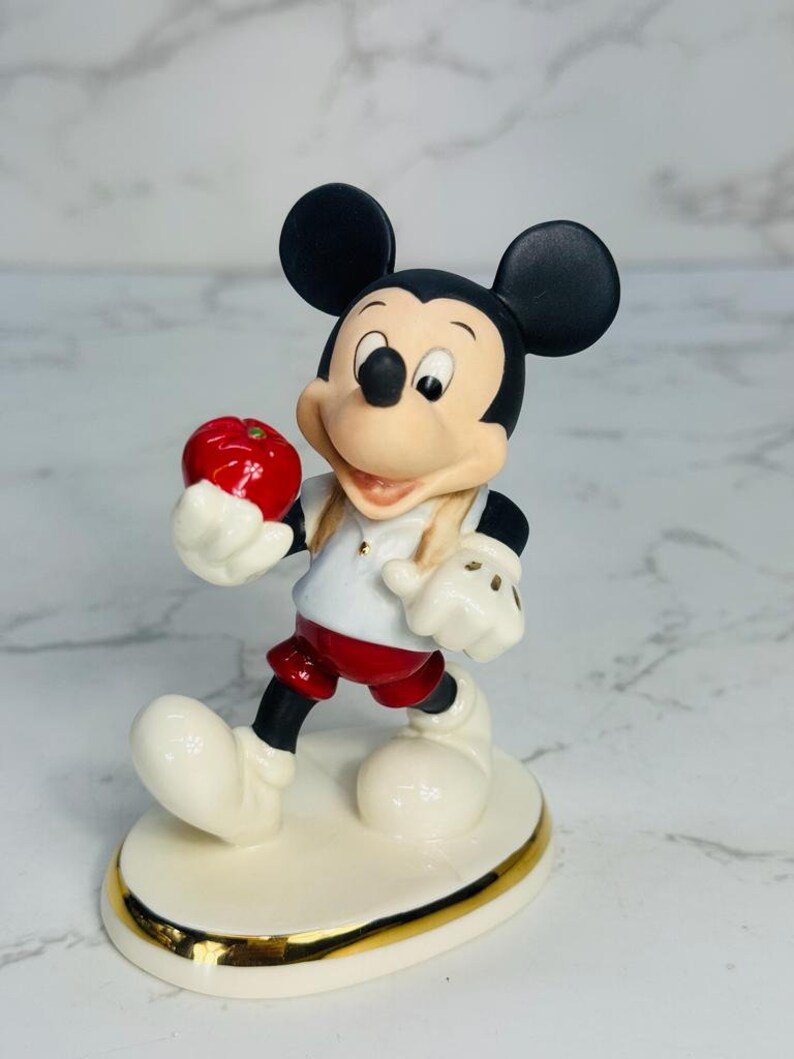 May include: A porcelain figurine of Mickey Mouse holding a red apple. He is wearing a white shirt with a red collar, red shorts, and white gloves. The figurine is standing on a white base with a gold trim.