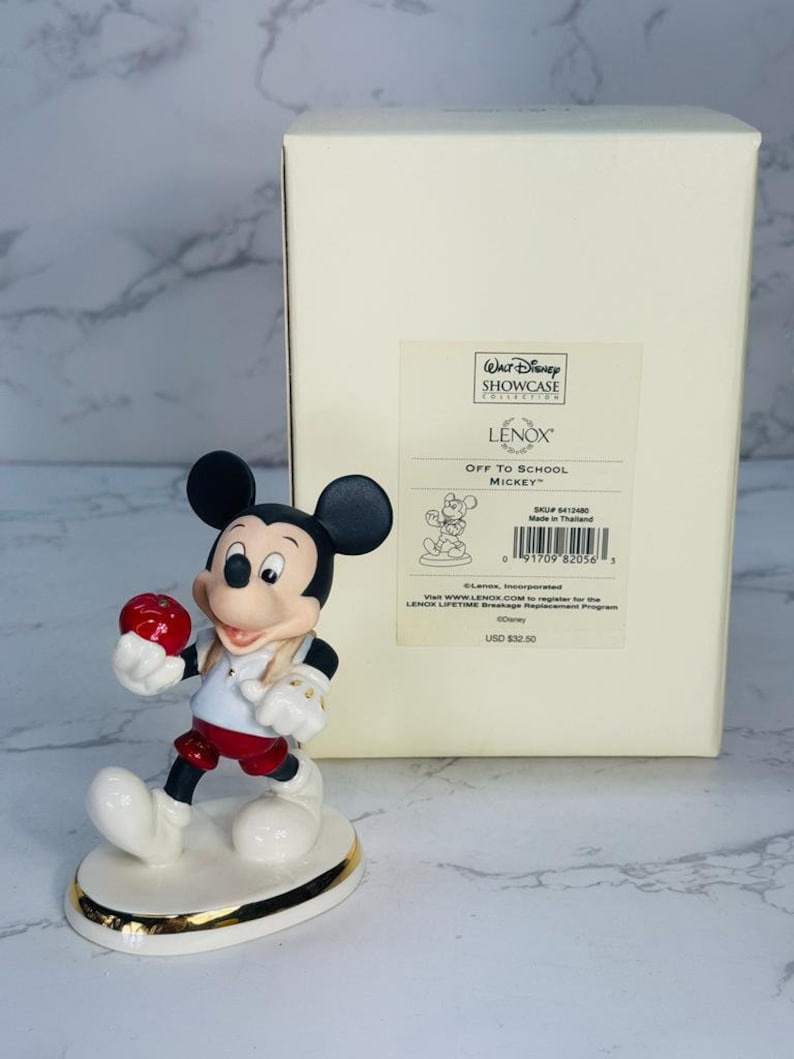 May include: A white porcelain figurine of Mickey Mouse holding a red apple. He is wearing a red and white shirt, red shorts, and black shoes. The figurine is on a white base with a gold trim. The box is white with the words "Walt Disney Showcase Collection" and "Lenox" printed on it. The box also has the words "Off to School Mickey" and "USD $32.50" printed on it.