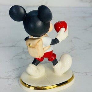 May include: A porcelain figurine of Mickey Mouse wearing a red shirt, red shorts, and a brown backpack. He is holding a red apple in his right hand and walking forward. The figurine is on a white base with a gold trim.