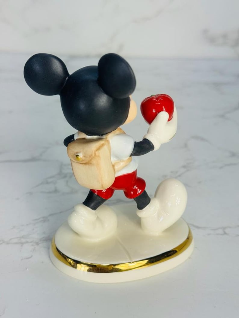 May include: A porcelain figurine of Mickey Mouse wearing a red shirt, red shorts, and a brown backpack. He is holding a red apple in his right hand and walking forward. The figurine is on a white base with a gold trim.
