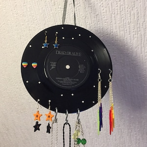 May include: A black vinyl record with white dots, used as a jewelry organizer. It has earrings, necklaces, and a chain hanging from it. The record label says "Dead or Alive" and "Lover Come Back to Me".
